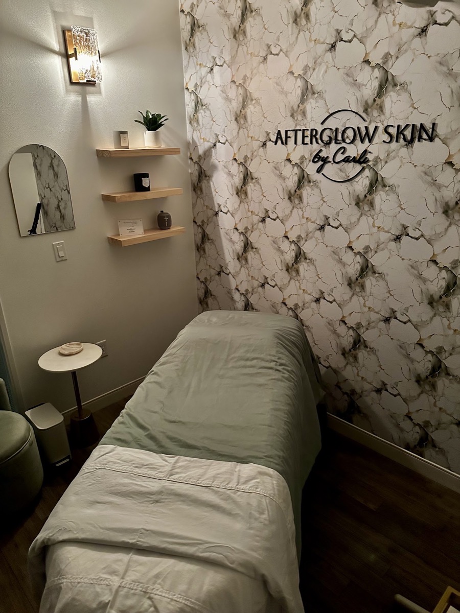 The AfterGlow Skin by Carli treatment room: a facial bed with sage-green linens beneath the studio&rsquo;s marble logo wall